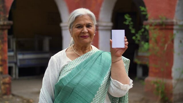 Grey hair Indian female happily showing her voter identification card before casting her vote in elections. Educated and responsible old lady all ready in traditional wear ready to give vote outsid...
