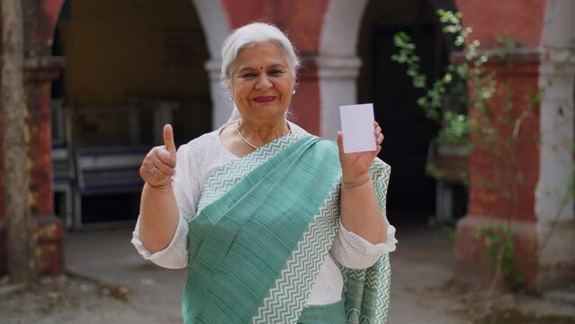 Grey-haired old lady dressed in saree  feeling happy after casting her vote in Indian elections - mature citizen. Smart old female showing her voter identification card while doing a gesture of thu...