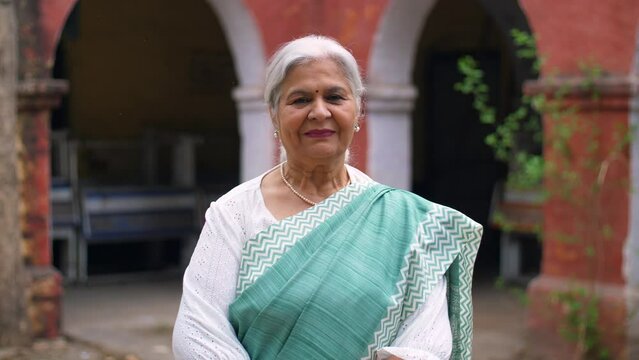 Grey hair Indian lady dressed decently in a saree posing and smiling on the camera - grand mother  mother-in-law. Old age businesswoman proudly standing outside an old building giving a toothy smil...