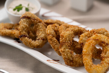 Deep fried calamari rings with sauce bowl serving on white plate in restaurant.