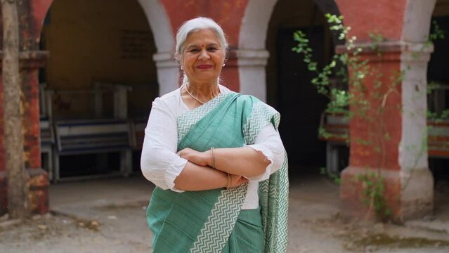 Old age Indian grand mother with grey hair posing or the camera while wearing a saree - senior citizen. Elderly woman standing confidently with a smile to pose for the camera with crossed hands - m...