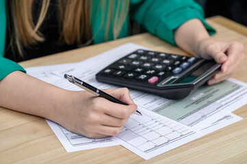 Kobieta siedzi przy biurku, wypełnia deklarację podatkowa i liczy na kalkulatorze  - obrazy, fototapety, plakaty