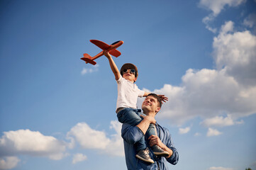 Playing with toy plane, sitting on the shoulders. Father and little son are having fun outdoors