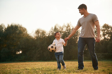 Fototapeta premium Walking and holding soccer ball. Father and little son are playing and having fun outdoors