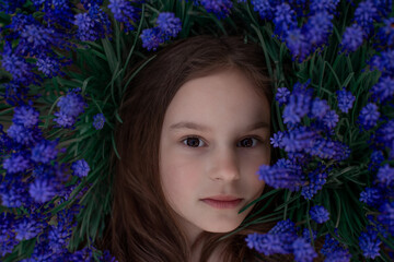 Portrait of young beautiful girl woman with brown hair lying on grass with blue flowers around her head. Close up, top view. Concept of spring summer. - 752259293