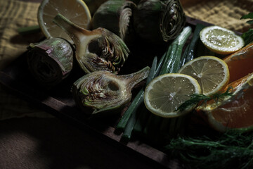 fresh artichoke flower buds with lemons, orange, green onion, herbs on wooden rustic board, yellow table cloth kitchen textile, vegetables ready to cook