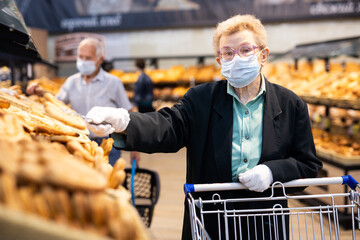 Older woman with glasses chooses buns and bread in supermarket bakery