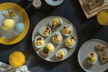 Happy Easter food background. Deviled eggs, orange juice, healthy crackers, dyed eggs on dark gray background with sunligh and harsh shadows, directly above - 752297209