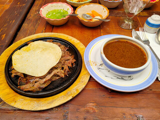 Juicy dish called birria served with rich soup and a handmade tortilla on a rustic wooden table