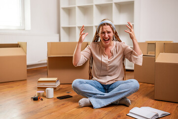 Angry ginger woman with braids shouting while sitting on the floor in her new apartment. She is...
