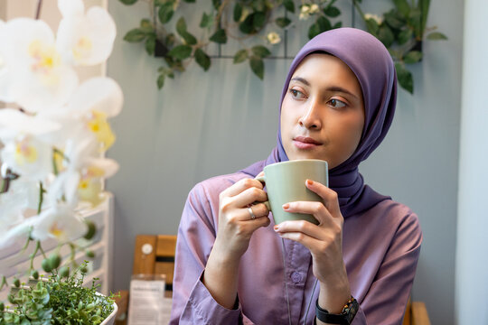 Beautiful woman in hijab sitting on balcony holding green army glass looking right, close-up front view, daydreaming or thinking concept