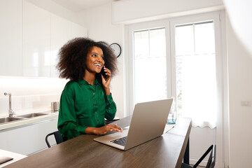 A joyful young woman with afro hairstyle is on her phone calling while working with laptop at her...