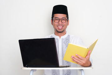 Moslem man sitting in front of laptop and holding a book showing happy face