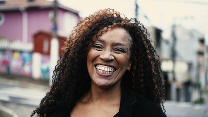 One joyful black woman smiling at camera in urban city environment in backdrop. Portrait close-up face of a 40s African American latin person - obrazy, fototapety, plakaty