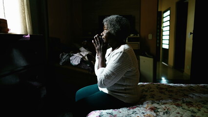 One faithful senior black woman seated by bedside Praying to God, gazing at window inside moody bedroom alone. Religious Elderly African American lady with gray hair