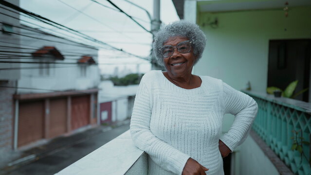 One happy elderly black woman from South America standing at humble residence balcony overlooking urban street in background. Portrait of 80s senior person with gray hair of African descent - Powered by Adobe