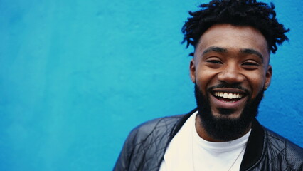 Obraz premium One young black man smiling and laughing on blue backdrop standing in city street. Portrait of an African American 20s person feeling happy