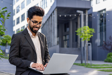 A smartly-dressed professional man engages with a laptop while seated outside a contemporary office...