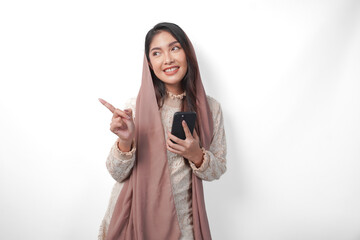 Happy Asian Muslim woman in veil hijab holding a smart phone while pointing to the copy space on...