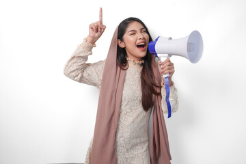 Young Asian muslim woman wearing headscarf veil hijab shouting at megaphone while pointing up to the copy space upwards, isolated on white background studio. Ramadan and Eid Mubarak concept.