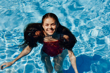 Happy woman swimming in pool in red swimsuit with loose long hair in sunshine, skin protection with sunscreen, concept of relaxing on vacation in tropical climate.