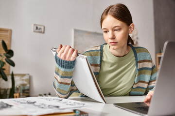 Focused teen girl looking at notebook while e-learning near laptop and sketches on desk