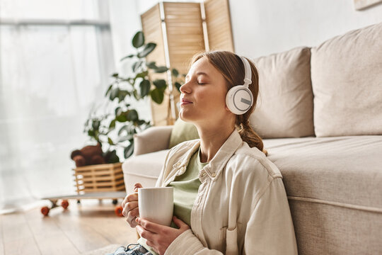 carefree teenager girl in wireless headphones listening music and holding cup of tea at home