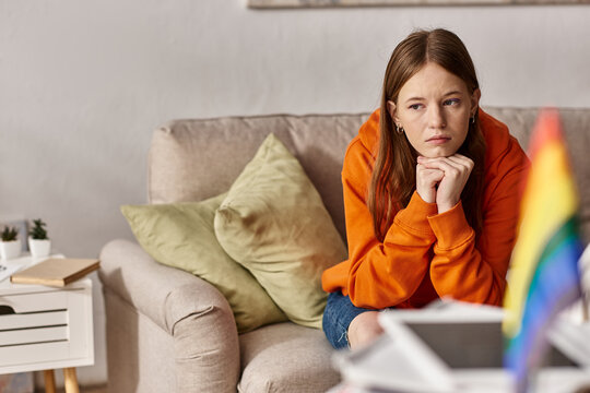 Pensive teen girl in hoodie sits on couch with a distant look, blurred lgbtq flag on foreground - Powered by Adobe