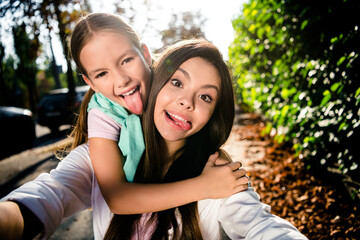 Naklejka premium Photo of two crazy cheerful sisters cuddle showing tongue out make selfie spend free time town park outdoors