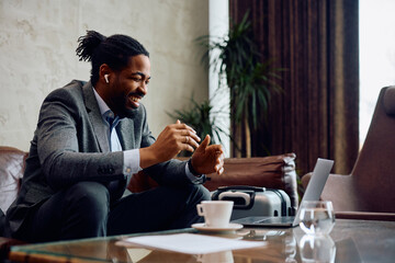 Cheerful black businessman having conference call via laptop in hotel lobby. - obrazy, fototapety, plakaty