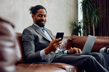 Happy black businessman texting on cell phone while working on laptop in hotel lobby.