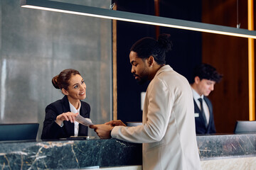 Happy receptionist assisting black male guest who is checking in at hotel.