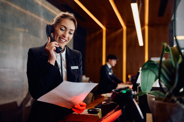 Happy receptionist talking on phone while going through paperwork at hotel front desk.