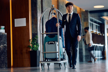 Happy bellboy pushing luggage cart with guest's suitcases in hotel.