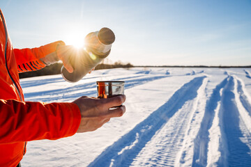 Man holding thermos with cup of tea, winter landscape, setting sun shining reflections on the...