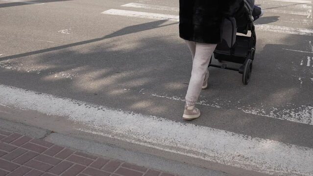 A young mother with a baby stroller crosses the road at a pedestrian crossing. A woman rolls her baby in a stroller across the road at a signalized intersection.