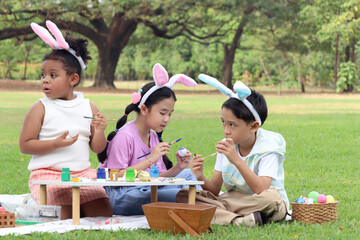 Happy three children in park, cute curly hair African girl with Asian boy and girl friend wear bunny ears, paint egg with paintbrush together in green garden. Kids celebrate Easter holiday outdoor.