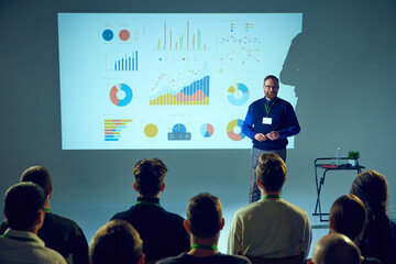 Presenter with ID badge stands in front of an audience, giving lecture with data chart backdrop in conference room. Concept of business, startup, leadership and personal development courses.
