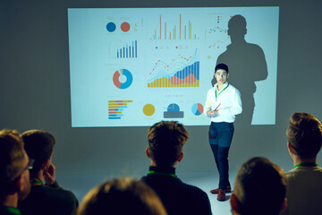 Asian man, businessman lecturing at business seminar with attendees focused on infographics displayed on screen behind him. Concept of business, startup, leadership and personal development courses. - obrazy, fototapety, plakaty