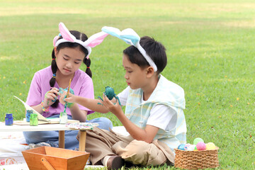 Cute Asian boy and girl with bunny ears painting egg with paintbrush while sitting together on...