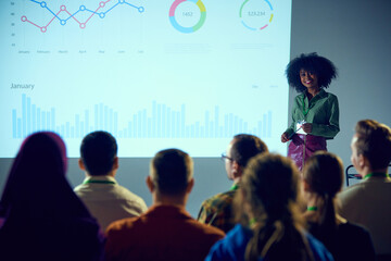 Young African-American woman, speaker at industry event presenting data analytics to group of...
