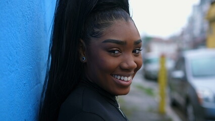 Portrait of a happy young black latina girl in 20s smiling at camera leaning on blue wall in urban...