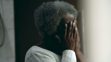 Regretful African American elderly 80s woman covering face with hands feeling anguish and worry about difficult circumstances during old age standing at home residence