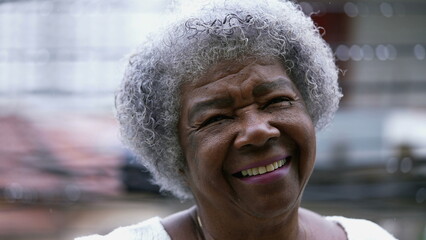 Portrait of a joyful black senior woman smiling at camera standing outside in drizzle. Carefree older African American person close-up face