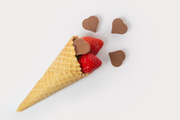 A waffle cone full of red ripe strawberries with green leaves and heart-shaped chocolates isolated on a pink background