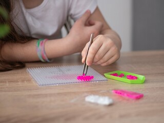 A close-up of a little girl's hand holding tweezers and assembling a toy from plastic parts of a thermo mosaic. The first stage of the work. Sensory education, hot plastic design.