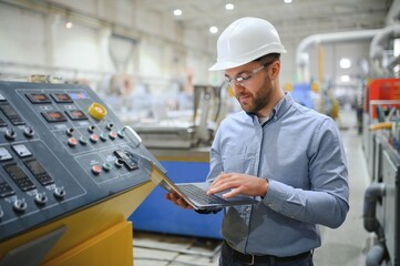 Engineer in hardhat is using a laptop in a heavy industry factory