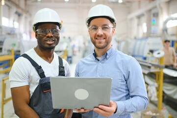 Two Diverse Professional Heavy Industry Engineers Wearing Safety Uniform and Hard Hats Working on Laptop