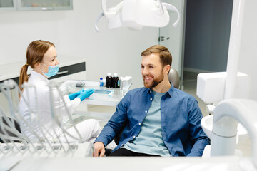 Blonde female dentist in dental office talking with male patient and preparing for treatment. Handsome bearded man in dentist chair looking at his doctor with smile