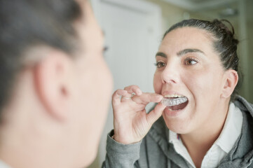 Brunette woman wearing invisible dental aligners looking into a mirror at her bathroom. Modern...